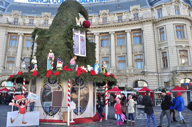 A lit Christmas tree and striped market stalls with visitors at Bucharest's Christmas Market, backed by a Belle-Époque facade