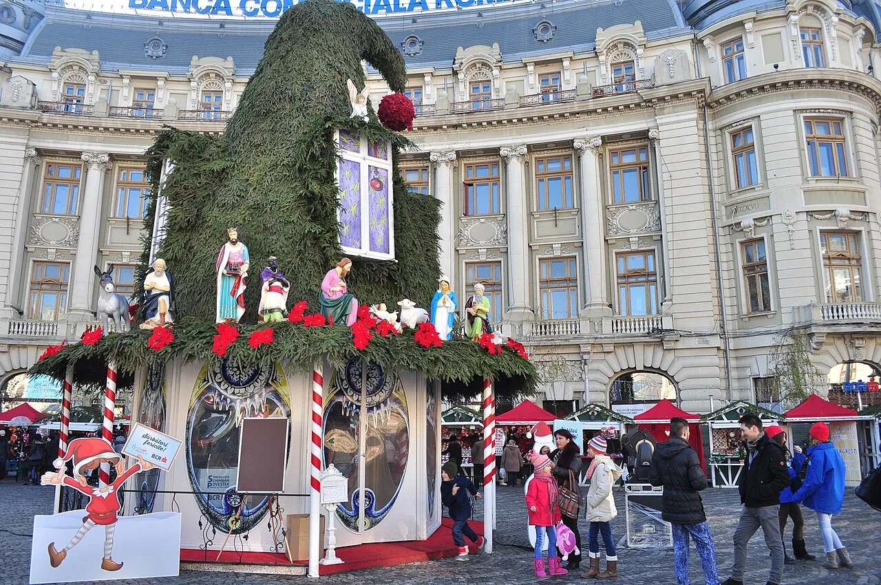A lit Christmas tree and striped market stalls with visitors at Bucharest's Christmas Market, backed by a Belle-Époque facade