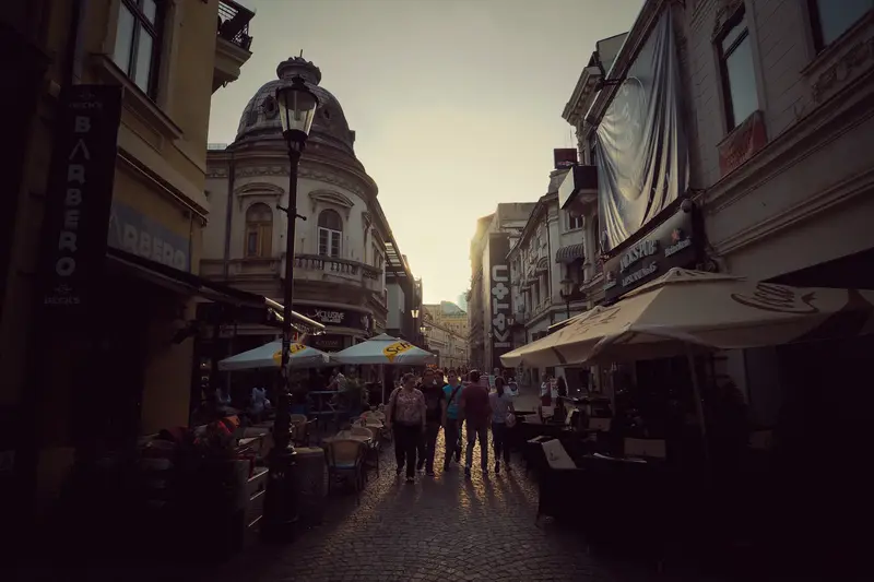 A lively Old Town street in Bucharest at golden-hour sunset, with people strolling among lit café and bar terraces