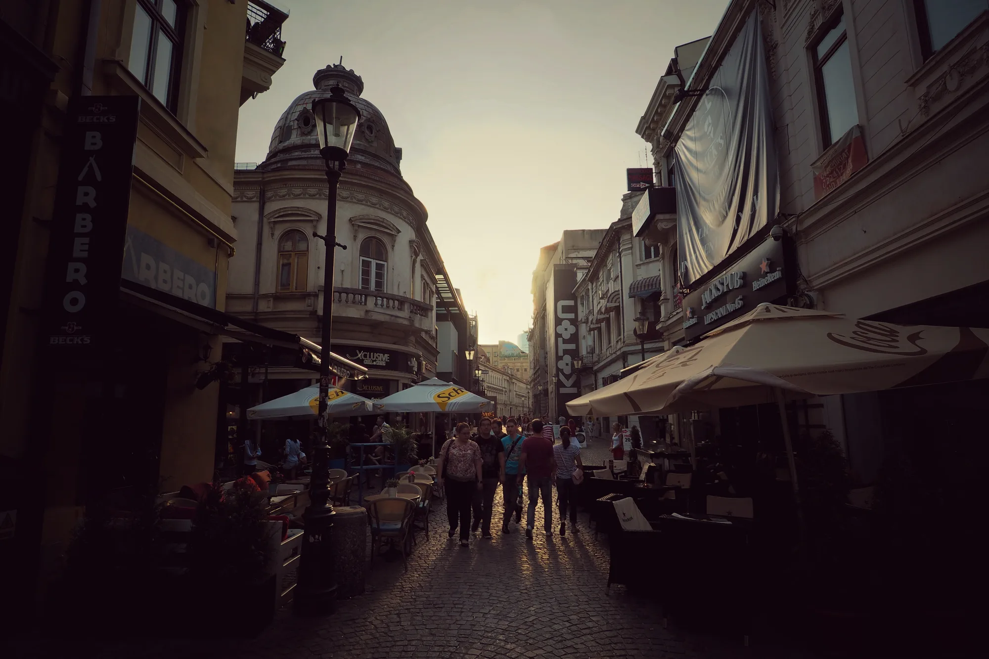A lively Old Town street in Bucharest at golden-hour sunset, with people strolling among lit café and bar terraces