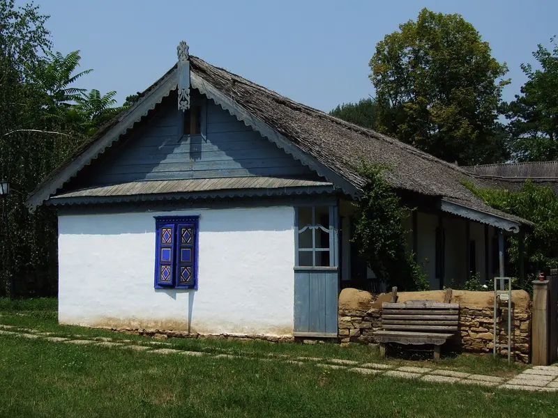 A traditional thatched-roof Romanian peasant farmhouse with whitewashed walls and blue woodwork at the open-air Village Museum in Bucharest